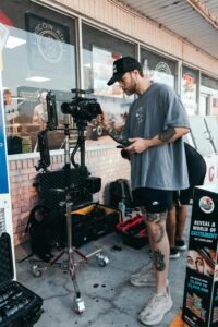 1. A tattooed man poses beside a camera, embodying the spirit of content creation and artistic expression. 2. A man adorned with tattoos stands next to a camera, representing the essence of modern content creation. 3. A tattooed individual stands beside a camera, highlighting the intersection of personal style and content creation.